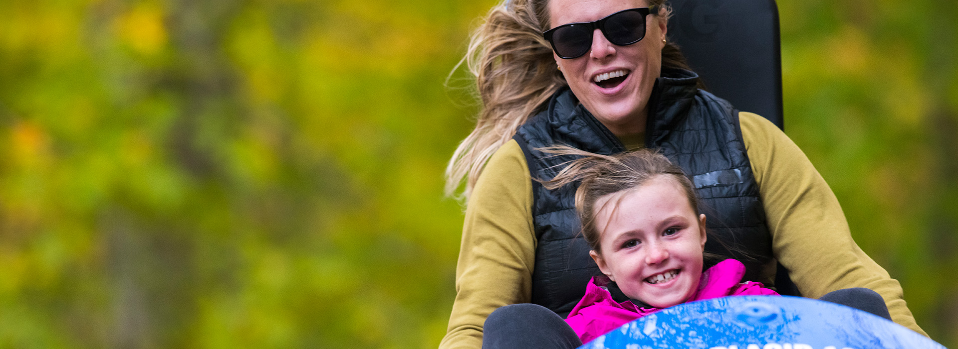 Woman and child smiling excitedly as they ride down the Cliffside Coaster at Mt Van Hoevenberg with the green trees in the background