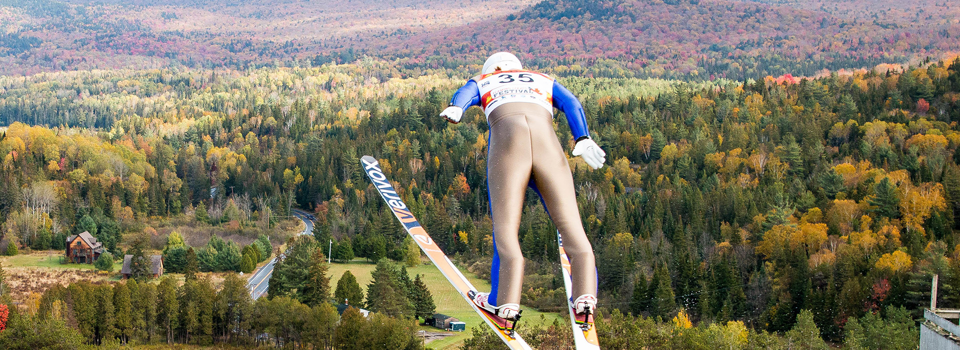 ski jumping fills much of the frame as he soars through the air high above the fall foliage in the background at the Olympic Jumping Complex.