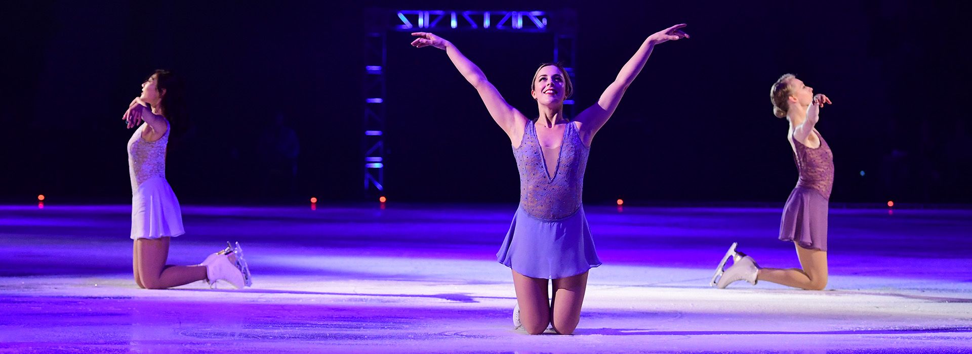 Female figure skater kneels on ice looking upward with hands in the air during a performance, at the Lake Placid Olympic Center