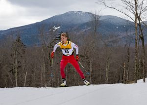 Nordic skate skier facing toward the camera climbs a hill at Mt Van Hoevenberg with Cascade Mountain in the background.