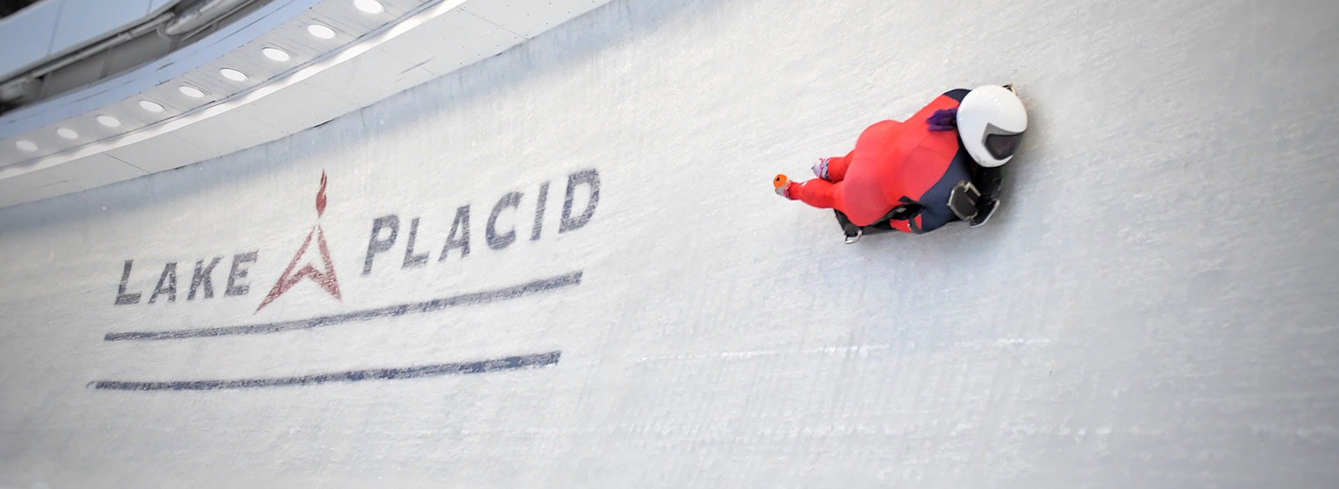 A skeleton athlete in a red unitard races around a turn with the Lake Placid Olympic logo emblazoned under the ice on the track.