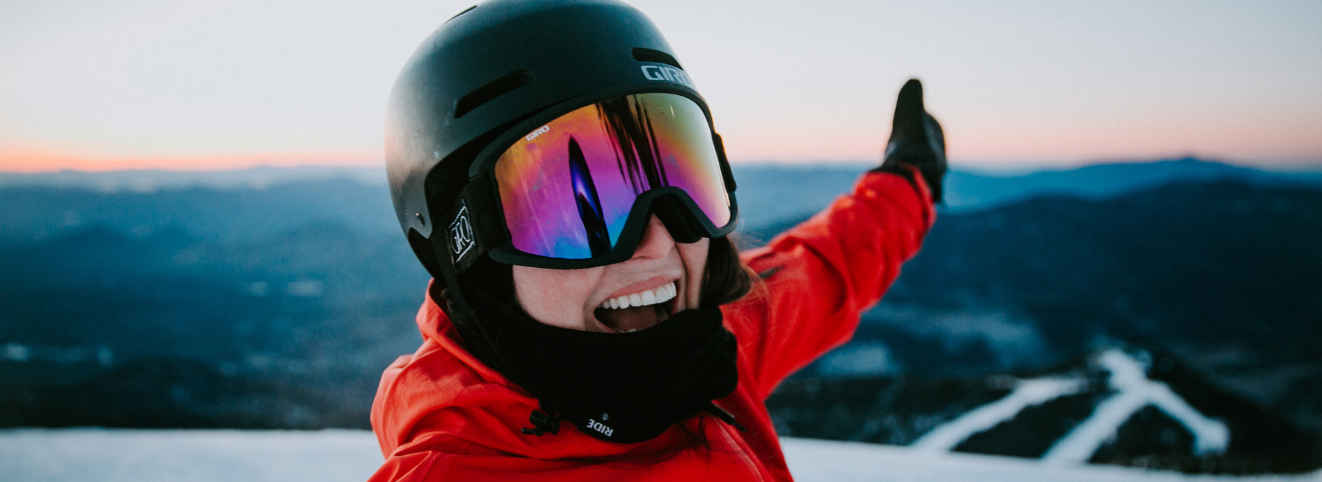 Woman in a helmet and goggles on a trail high on Whiteface Mountain with her arm in the air and a big open smile with excitement about the mountains and trails below her in the background.