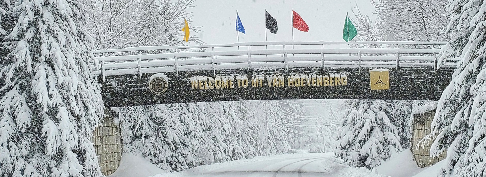 Snow covered road with bridge and sign that reads "Welcome to Mt Van Hoevenberg."