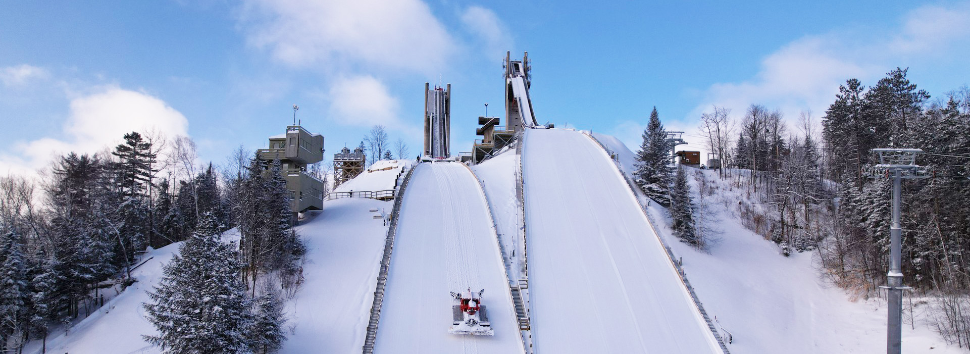 The Olympic Jumping Complex HS 100 meter and HS 128 meter jumps taken from the end runs below the towers on a bright, partly cloudy winter day with a snow groomer partway up the landing hill.