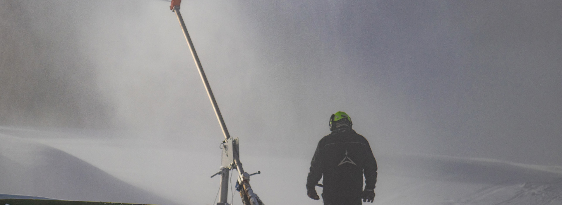 A lone snowmaker on the mountain enveloped in a white mist of snow particles with a metal snowgun at his feet sticking up into the air.