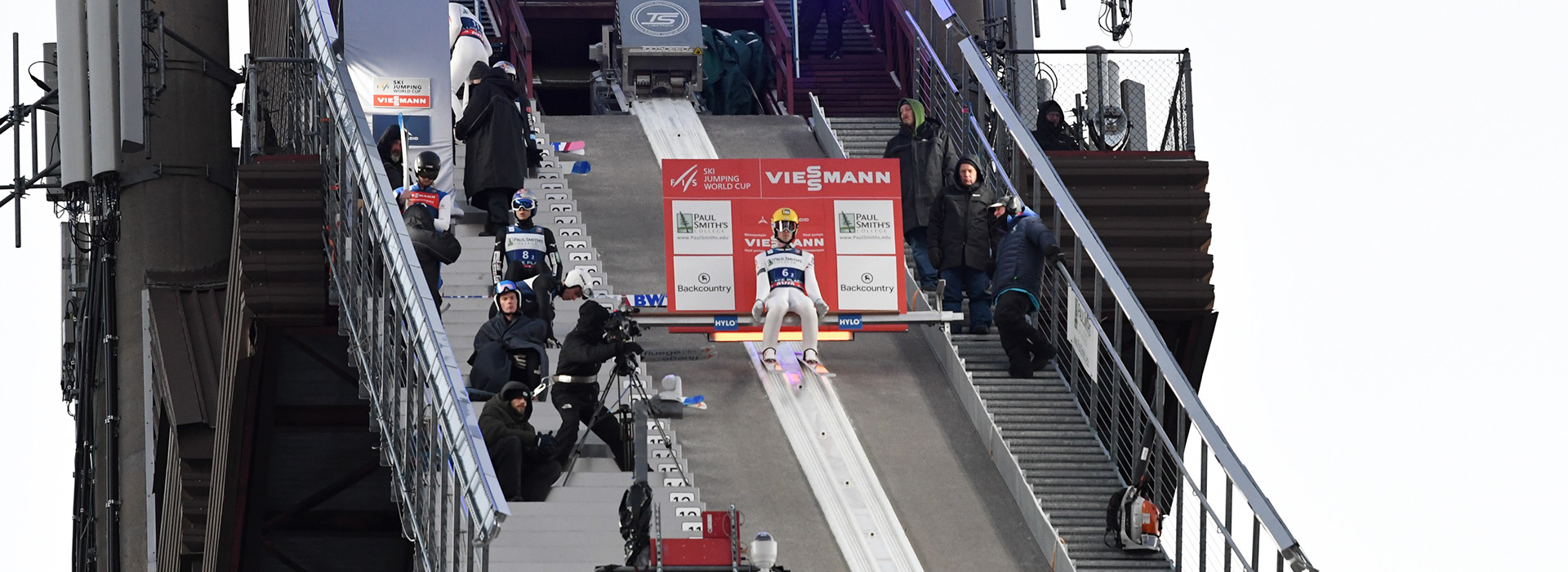 Image of a World Cup ski jumper sitting on the top of the inrun preparing to go with other athletes, staff, and volunteers on the stairs surrounding him.