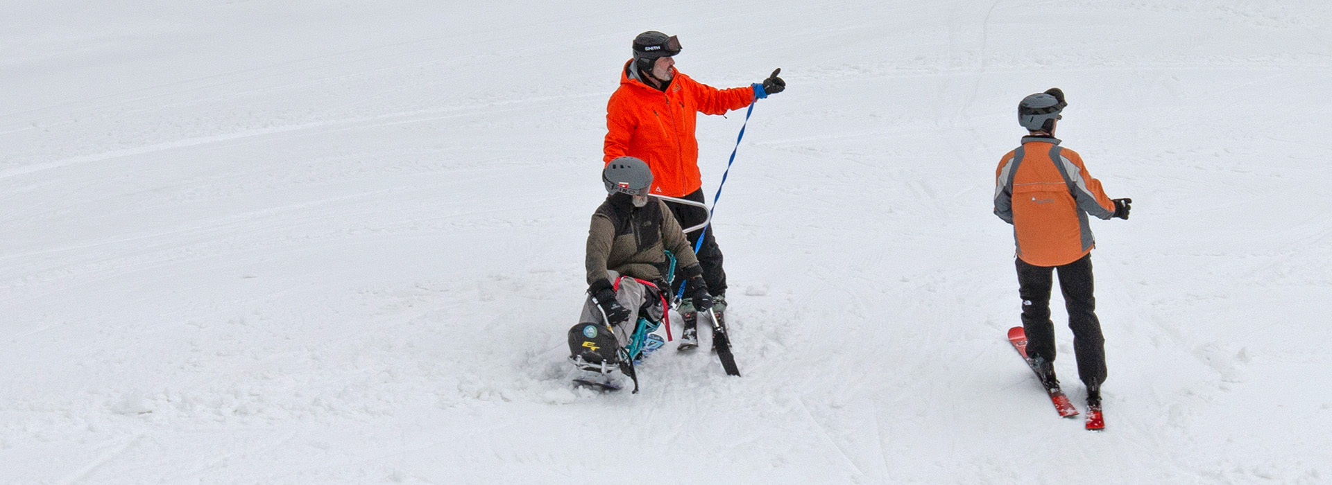 A ski instructor in an orange coat who is helping a disabled skier in a monoski points down a snow covered slope while another volunteer assistant looks on.