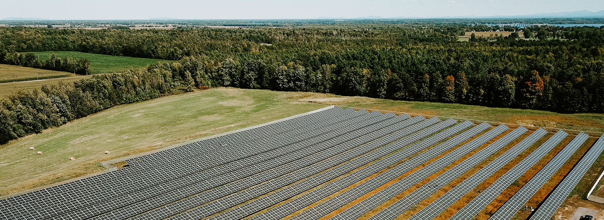 A portion of a large solar array at the bottom of this photo surrounded by field and forest with the horizon visible in the background.