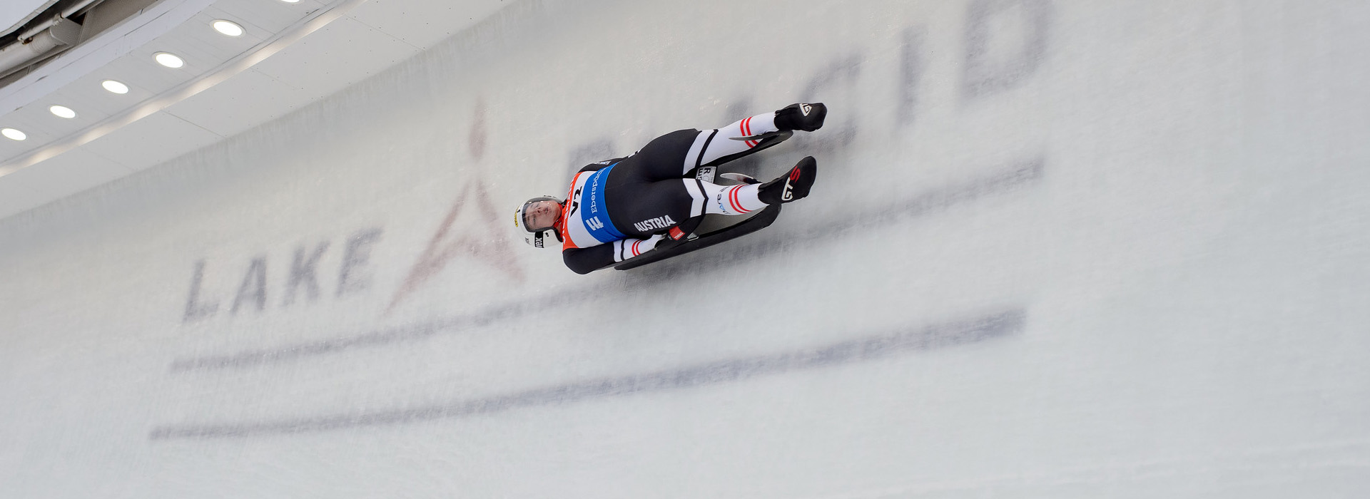 An Austrian male luge athlete making a turn high on the track with the Lake Placid logo appearing in the ice under him.