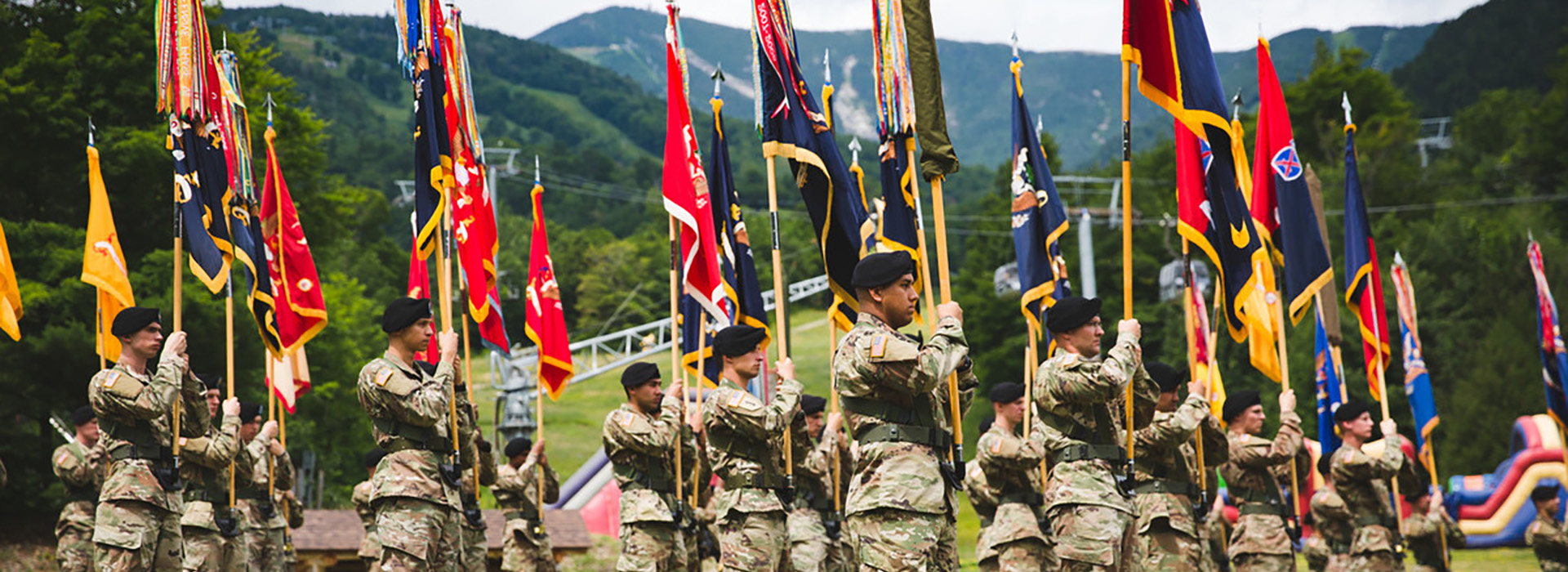 Lines of soldiers from Fort Drum dressed in camouflage uniforms and holding colorful flags stand solemnly at the base with trees and the Whiteface summit in the background