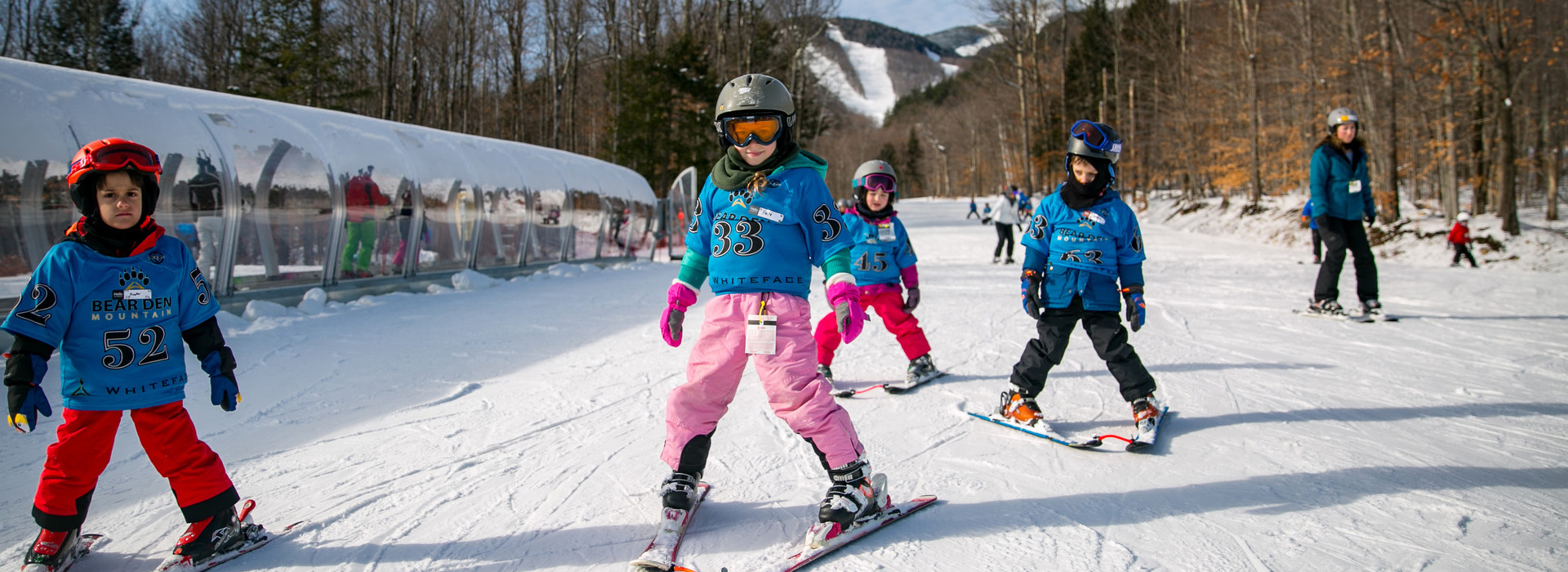 Young children on skis facing the camera and navigating a snowy surface next to the covered magic carpet lift at the Bear Den Learning Center.
