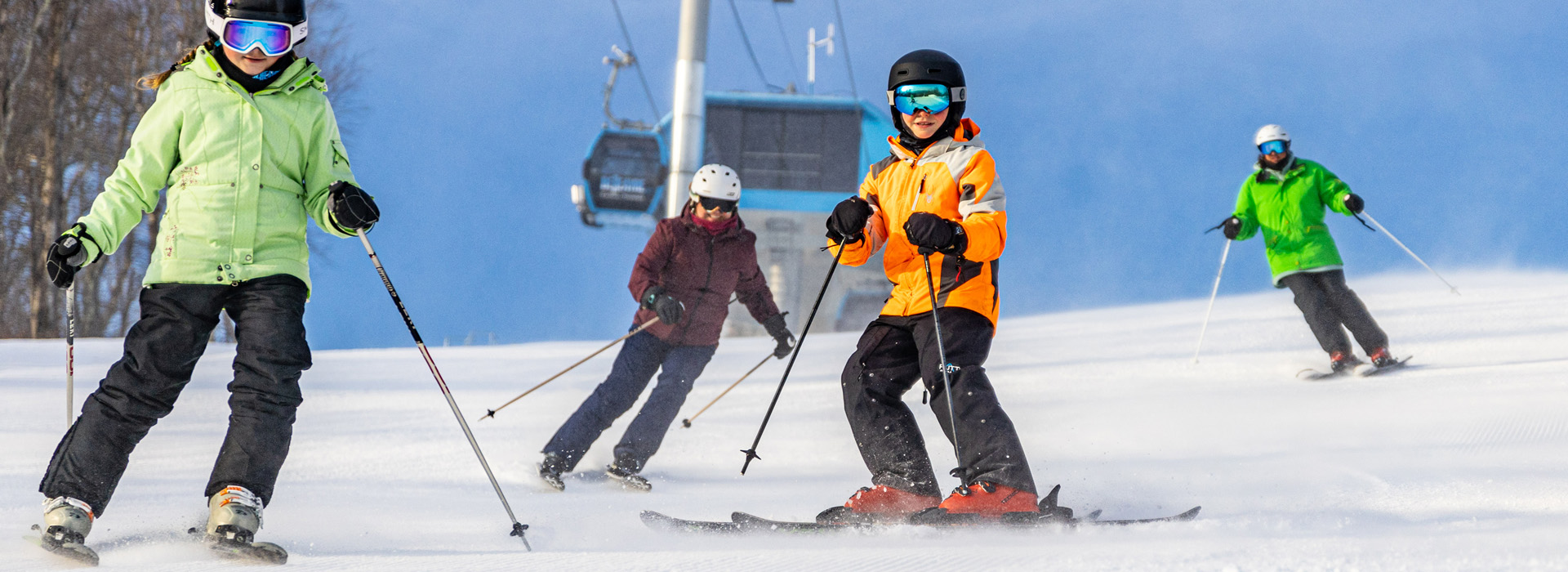 Four skiers in all, including two youth skiers in the foreground looking at the camera, all on the slope with the gondola and the summit in the background.