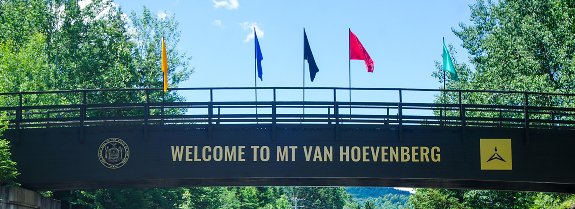 The bridge over the road entering Mt Van Hoevenberg is pictured in sukmmer with its colored flags and the words, "Welcome to Mt Van Hoevenberg"