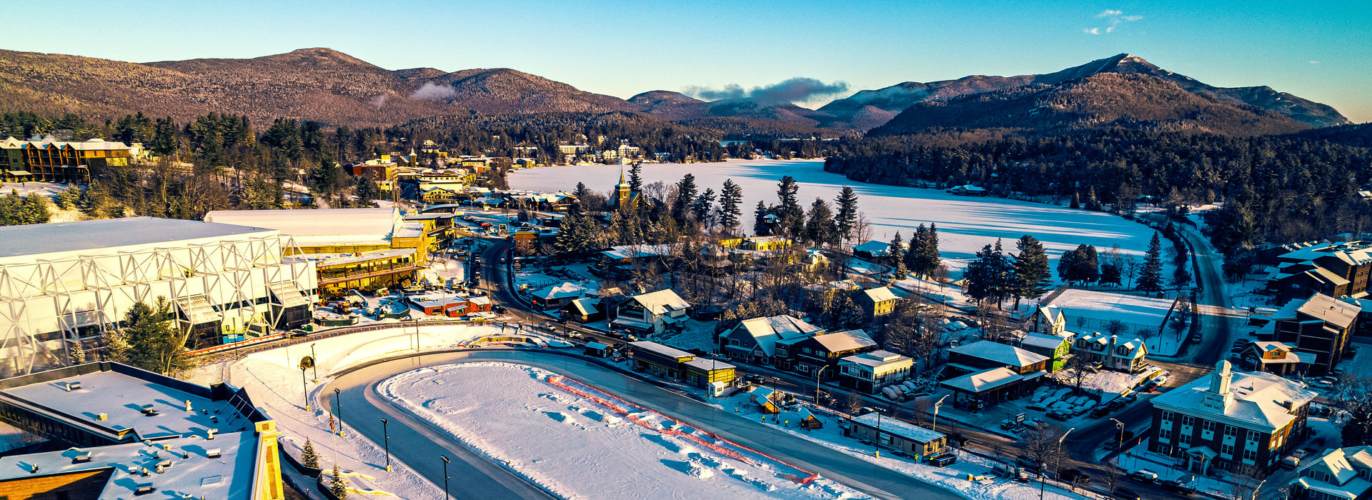 An aerial view of Lake Placid's Olympic Center and downtown region on a bright winter day, showing the oval in front and Mirror Lake and mountains in the background.