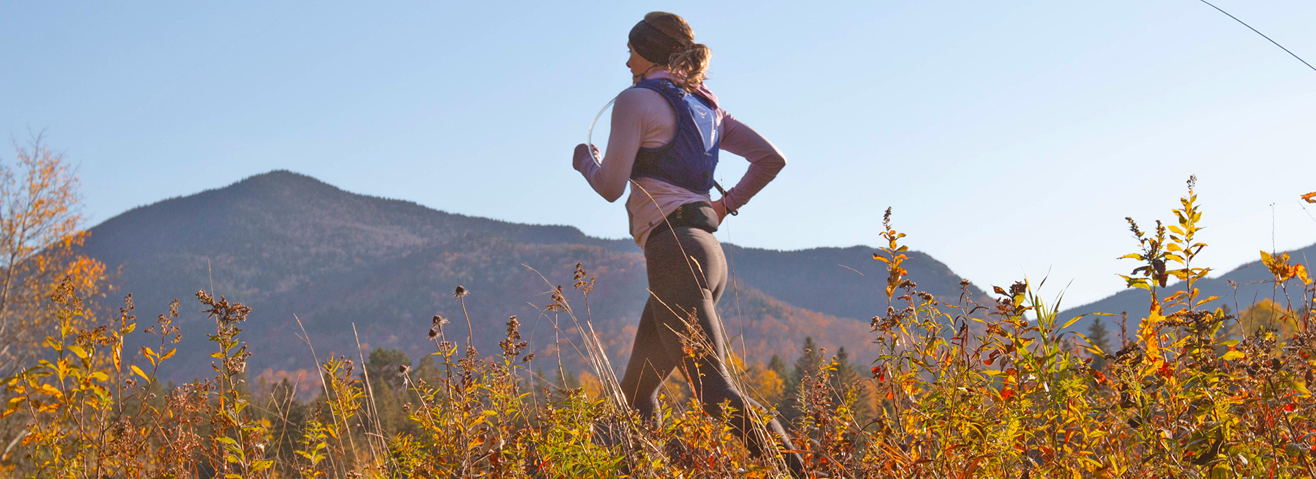 Woman with a hydration pack running through tall vegetation in the autumn at Mt Van Hoevenberg with views of the mountains in the background.