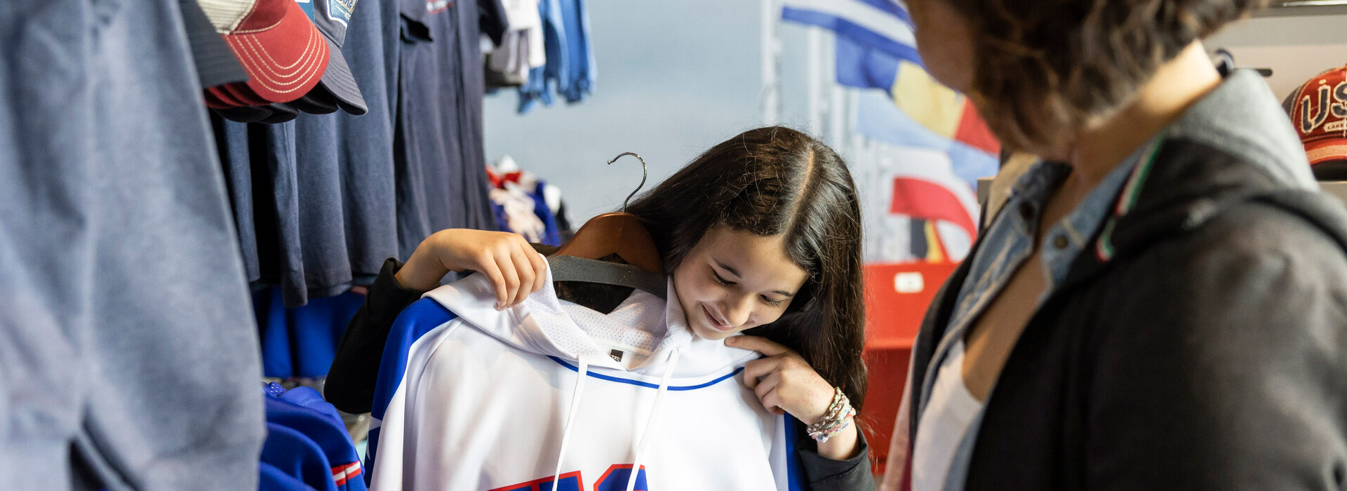 A young girl with dark hair holds up a USA branded hoodie against her as she contemplates a purchase with her mother on the right looking on.