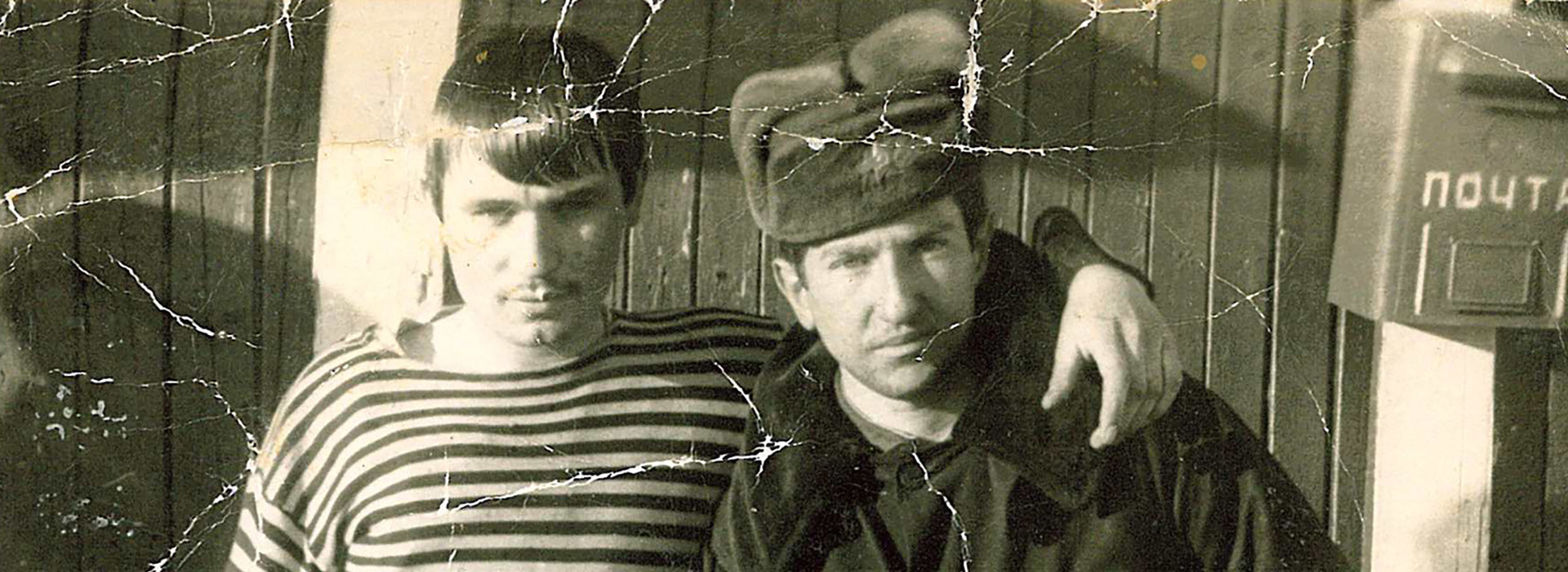 A black and white photo with wrinkle marks of a young Dmitry Feld in a fur hat in the former Soviet Union with another unidentified man in a striped shirt.