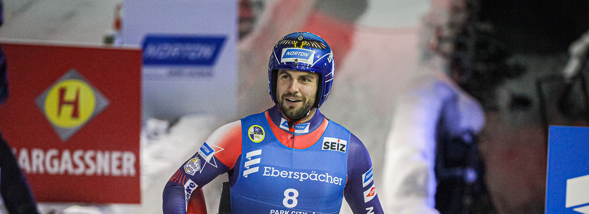 Portrait of Chris Mazder taken as he walks down the luge track after finishing, carrying his sled with his helmet on