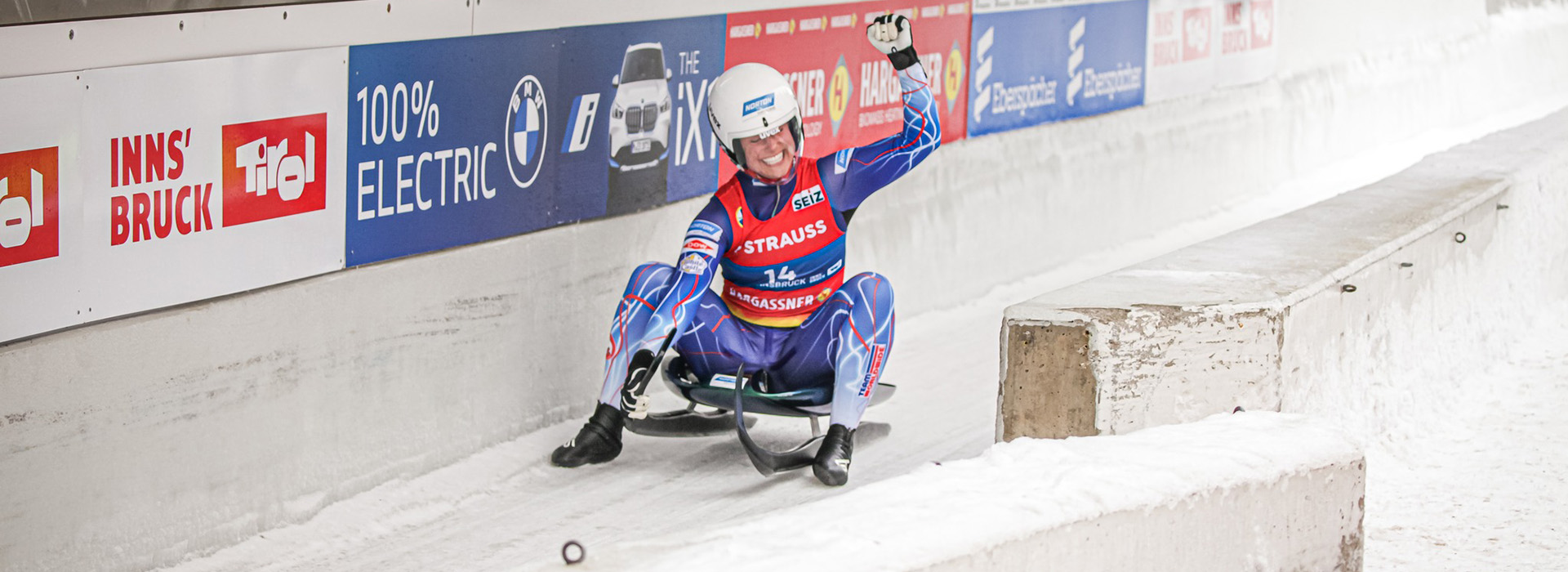 Emily Sweeney on her luge sled at the finish of a run with a big smile on her face and her hand raised in the air triumphantly.