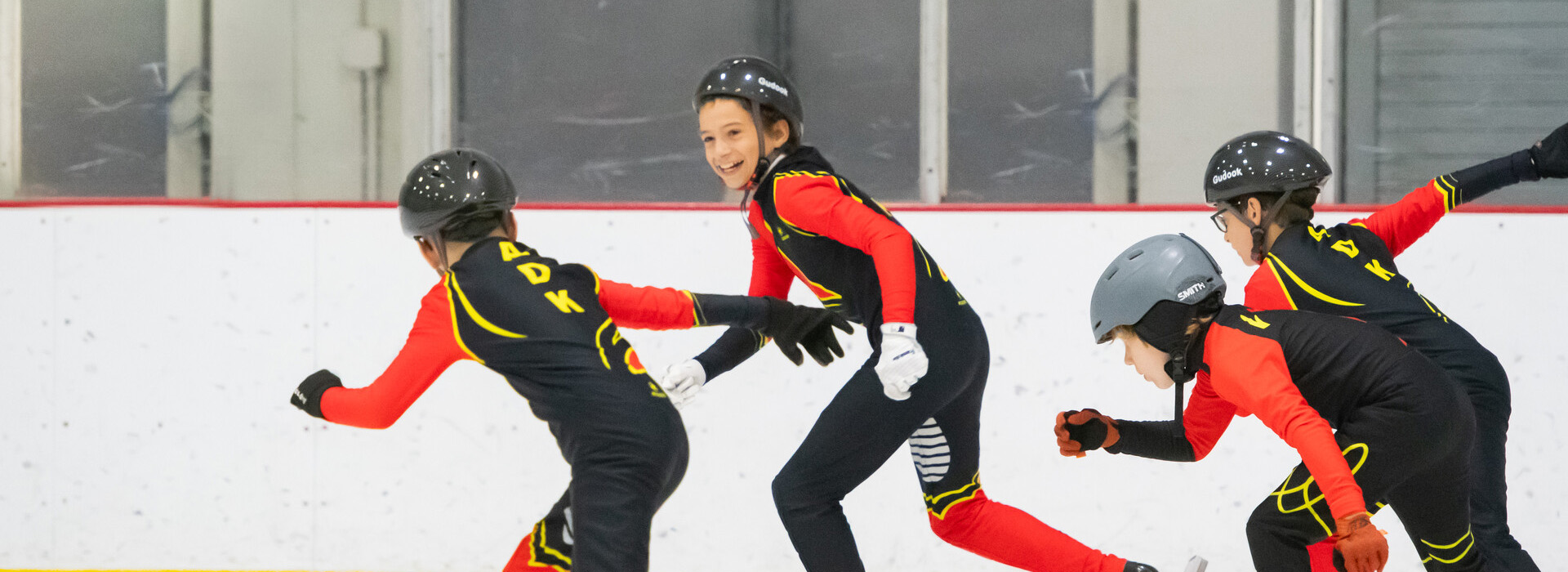 Several children in matching ADK Speedskating Club uniforms skating on an indoor rink, one of them looking at another and smiling big.