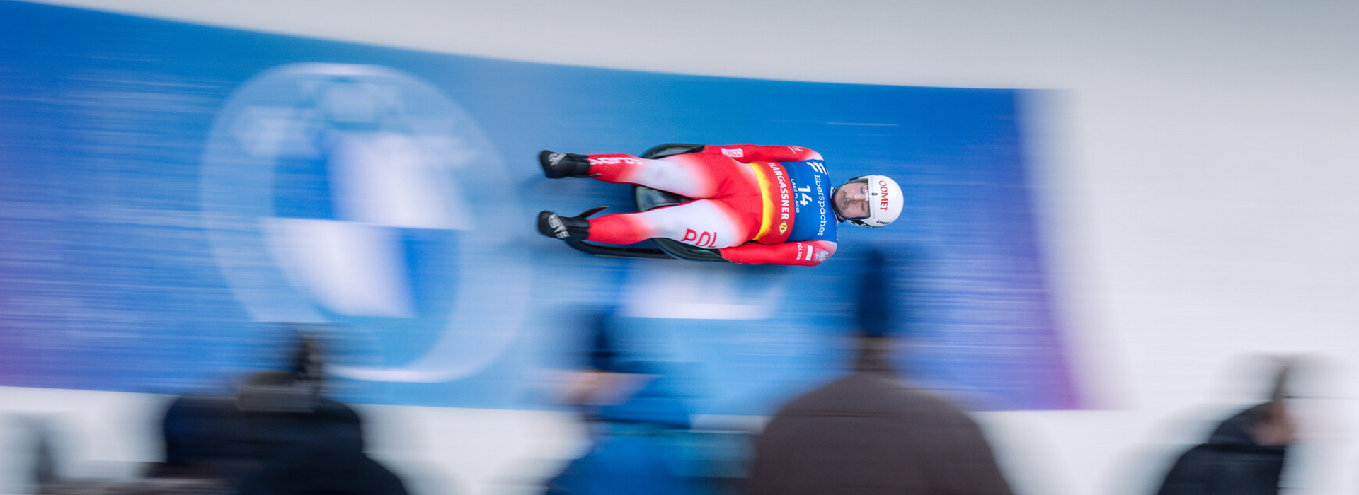 A luge athlete in sharp focus rushes high around a banked turn with spectators in front who are out of focus.