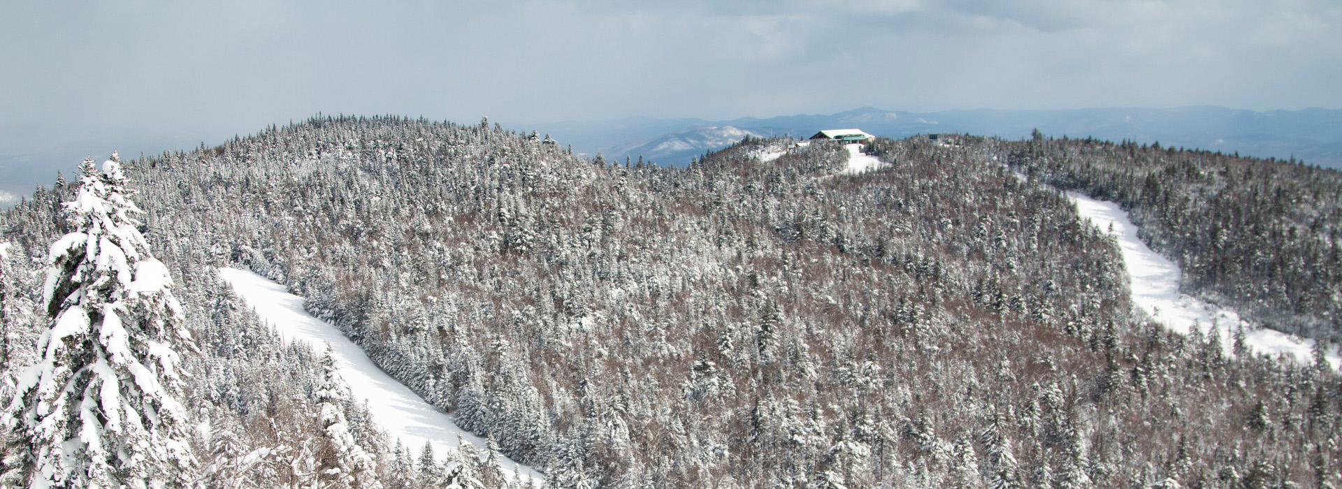 Gore Mountain landscape photo in winter with snow laden cloudy skies, a few ski trails amidst the forested mountains, and a small lodge on a peak.