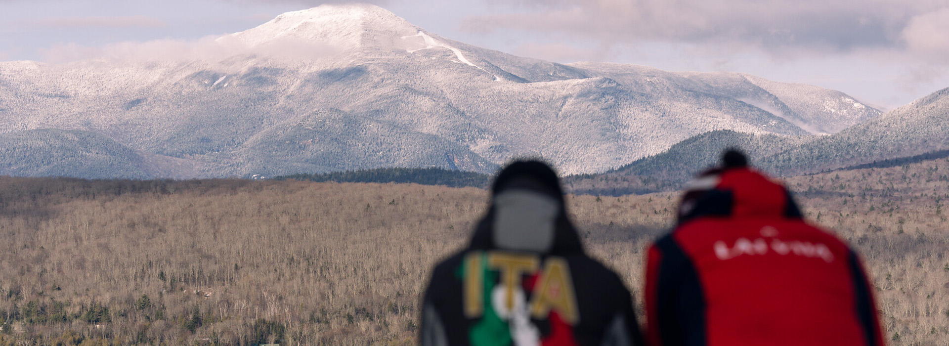 Winter view of Whiteface Mountain from Mt Van Hoevenberg with an athlete from Italy and another in a red jacket in the foreground looking on, with the athletes slightly out of focus.