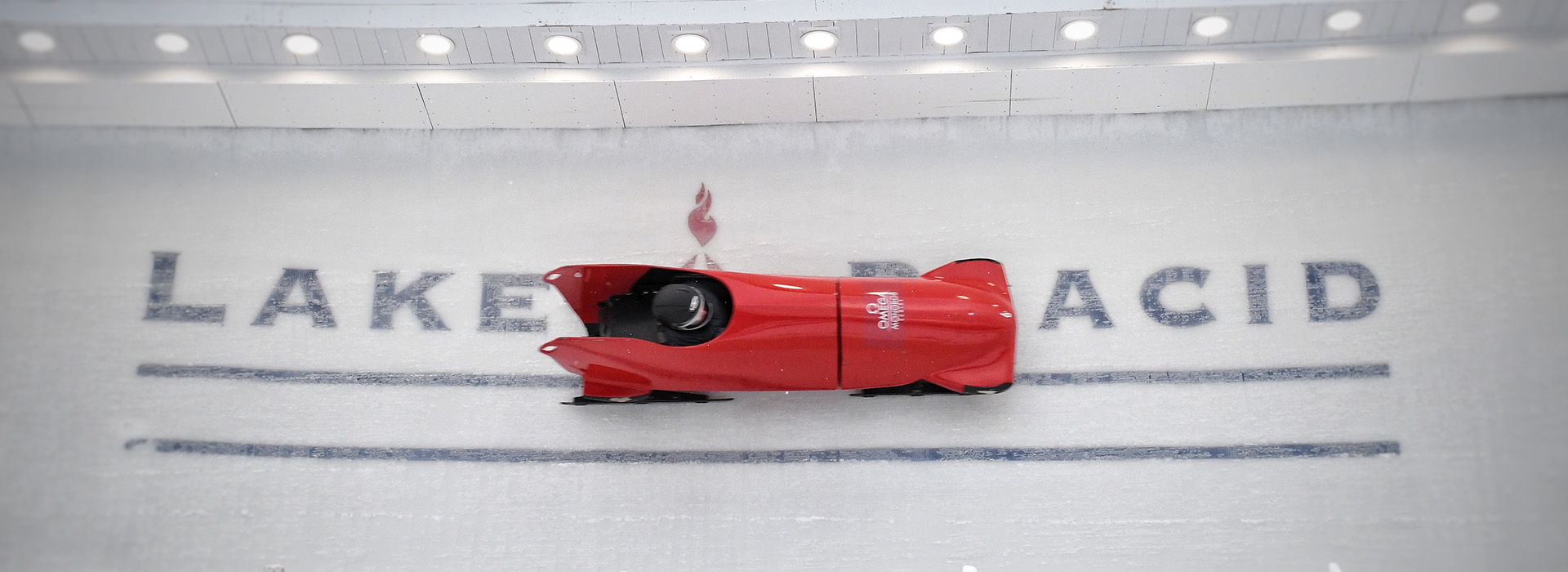 A red mono bob makes a turn on the combined track high on a wall during the IBSF Para Bobsled World Cup competition at Mt Van Hoevenberg with the Lake Placid logo appearing in the ice under the sled.