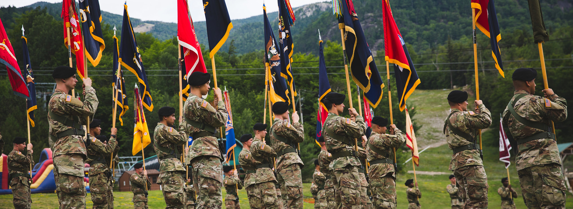 Soldiers from the 10th Mountain Division at Fort Drum at the Whiteface Mountain Rededication Ceremony in summer standing in formation wearing camouflage and holding flags with the green foliage of the mountain in the background.