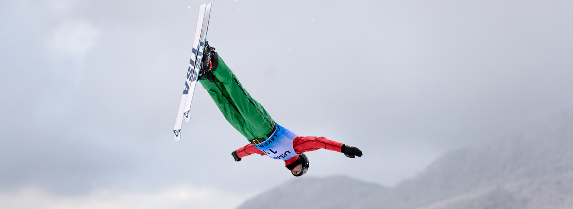 An aerials ski jumper in mid flight with green pants and a blue and black bib, red sleeves, inverted in the air against a cloudy gray background with the faint image of mountains in the distance lower right.