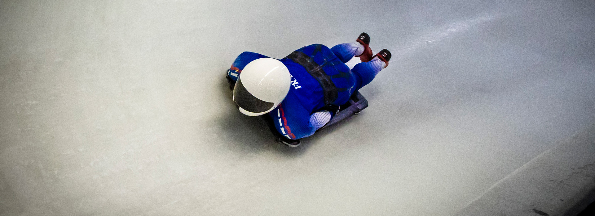 A skeleton athlete in a blue France uniform with a white helmet sliding down the Mt Van Hoevenberg track with nothing but ice showing around them.