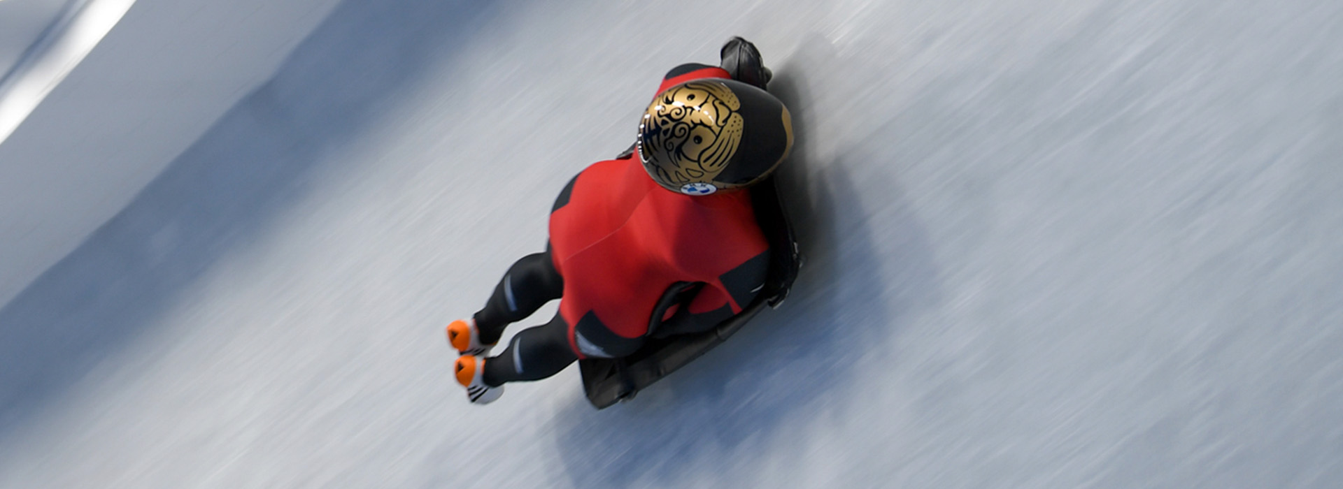 A skeleton athlete in red with a gold helmet with an intricate design is sliding around a steep turn, only the ice and the athlete visible in the photo.