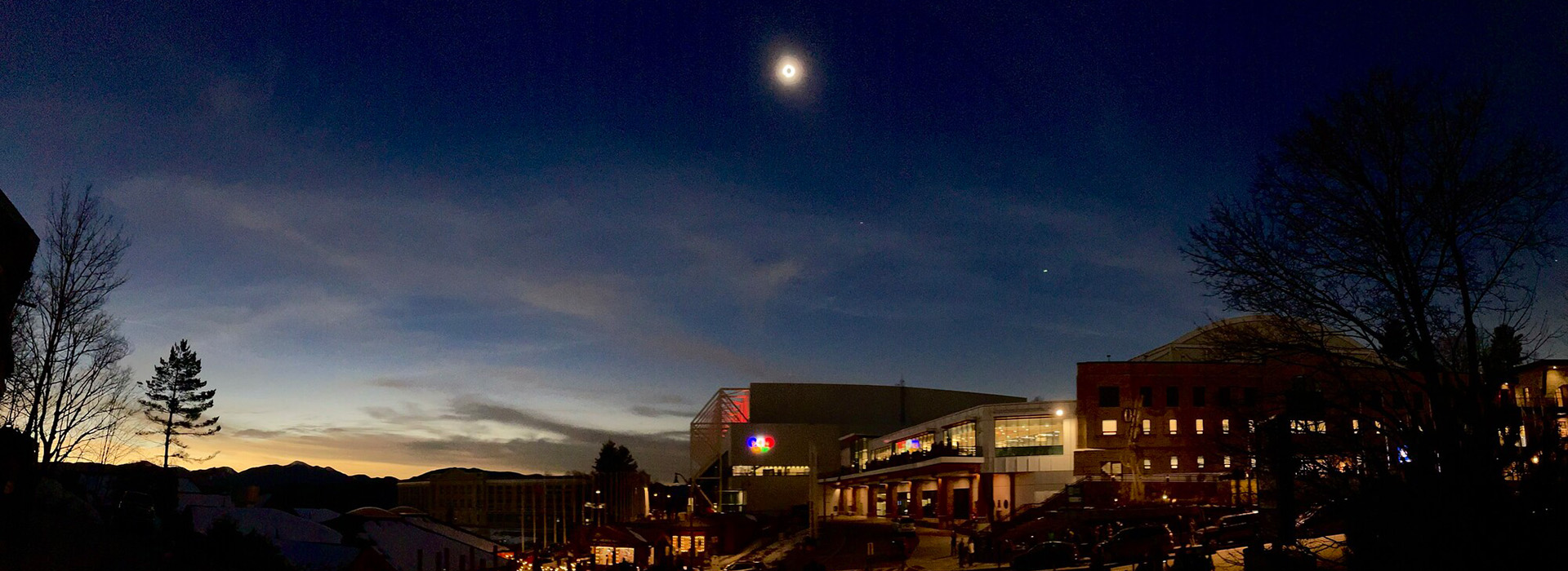 The total eclipse of the sun on April 8, 2024, high in the sky above the Olympic Center with the Olympic rings illuminated on the side of the building and internal Olympic Center lights showing a crowd on the deck and on the street.