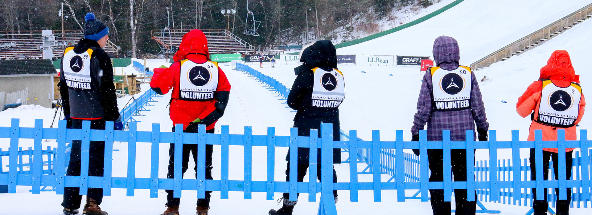 Five volunteers in winter coats with bibs that say "volunteer" on the back facing away from the camera outdoors with a blue fence in the foreground and cross country skiers and bleachers in the background.