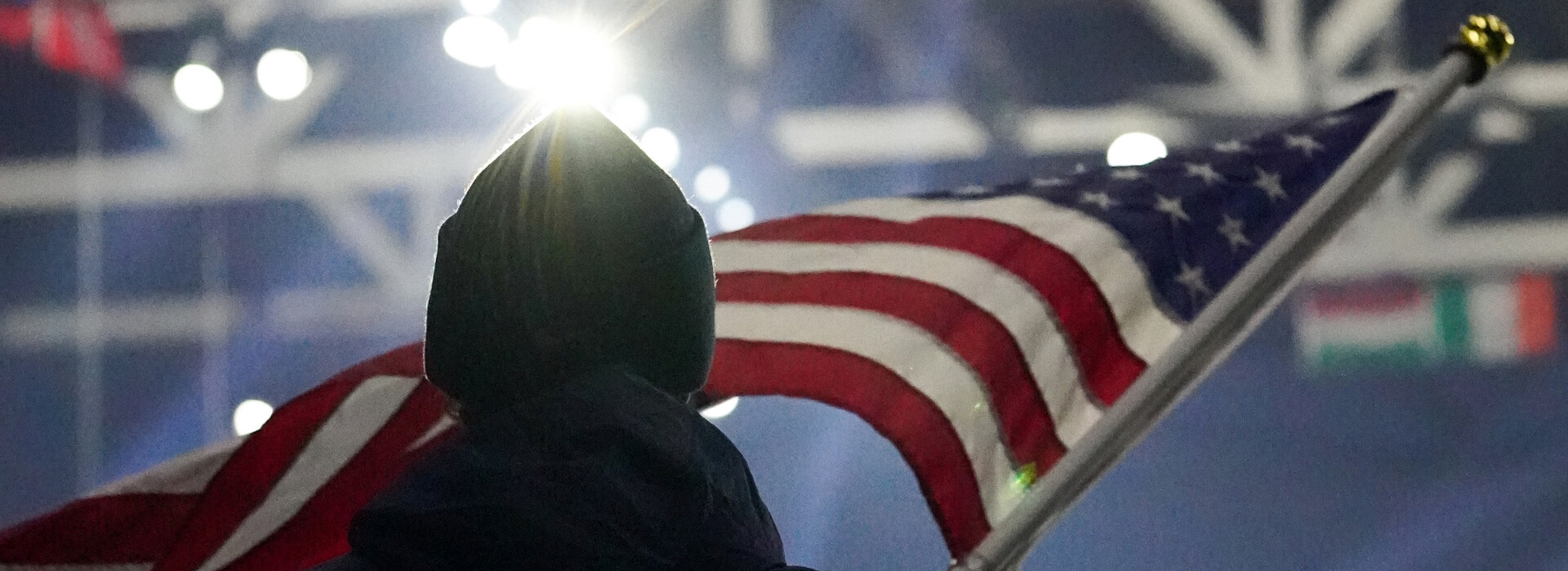 Image from behind a person with a dark stocking hat waving a U.S. flag during the opening ceremonies of the 2024 FISU Winter Games inside the Olympic Center with bright lights in the foreground.