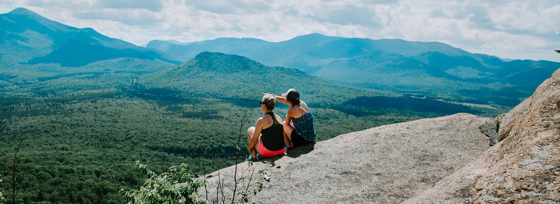 Two women sitting on the summit of Mt Van Hoevenberg is summer with one of them pointing into the mountains in tje distance.