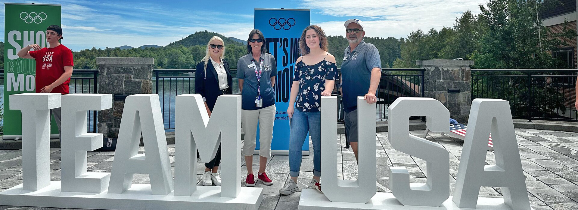 Olympic Authority staff at Mids Park in Lake Placid with Mirror Lake in the background and a Team USA word display in the foreground.