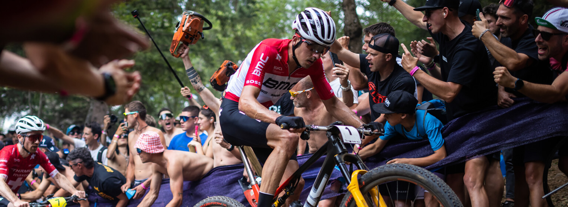 A male rider pedals uphill in a big mountain bike race with a crowd of spectators cheering and waving at the barrier.