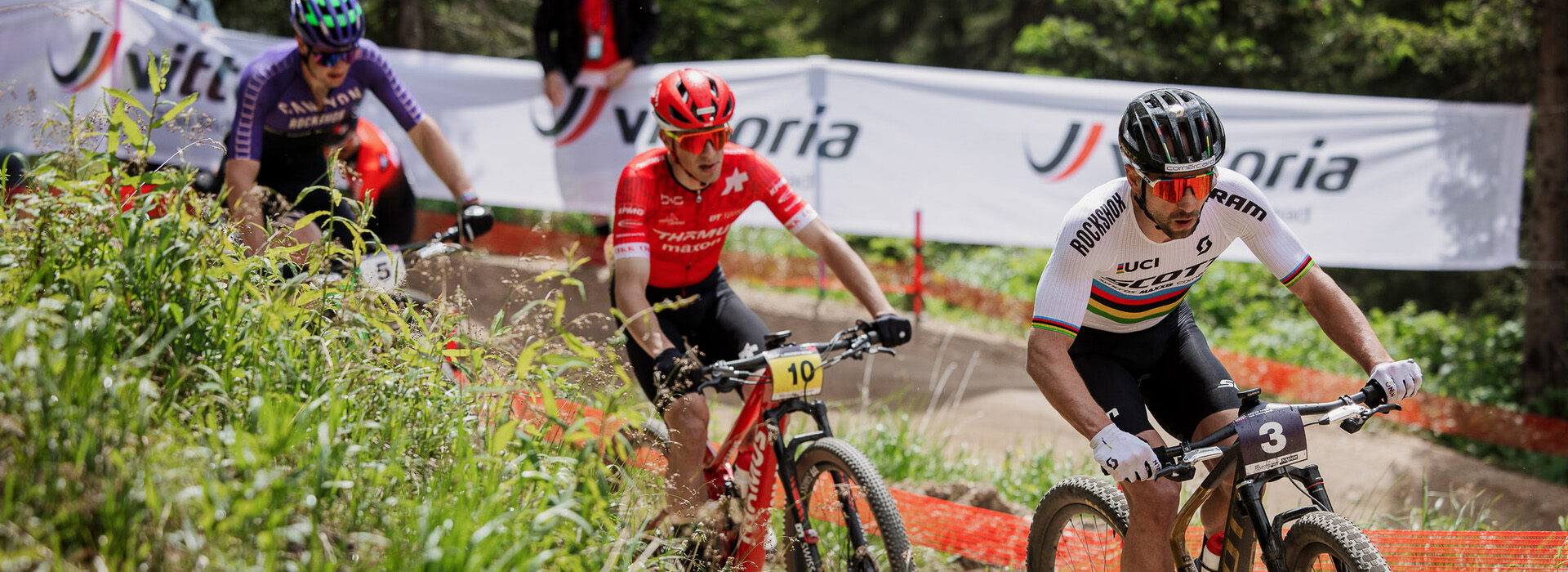 Three male mountain bike athletes riding single file on a trail during competition.