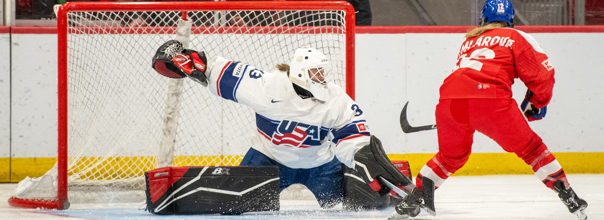 A female goalie in a mostly white uniform for Team USA stretches out to made a save during a game as the opponent in all red just in front of the net tries to score.
