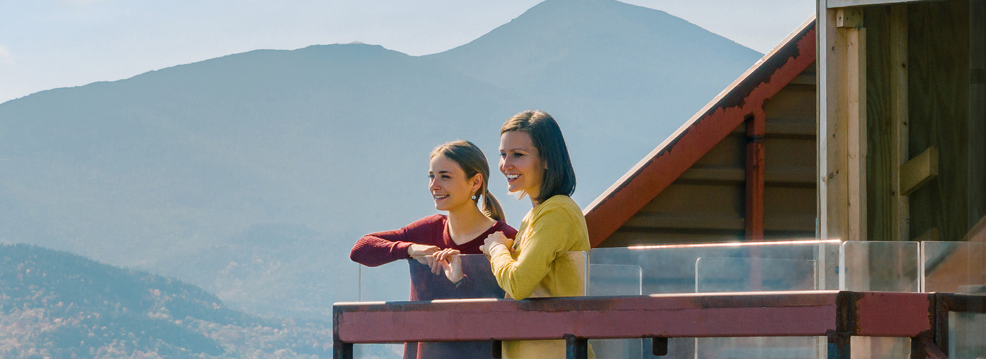A mother and daughter admire the view on a sunny fall day from the top of the Ski Jump platform with mountains in the background.