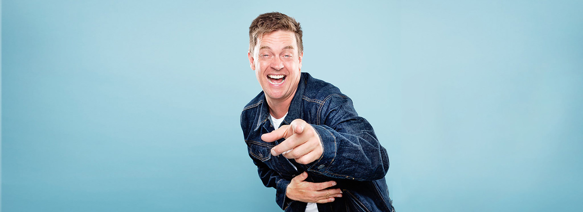 Portrait of comedian Jim Breuer against a light blue background with him laughing bent over and pointing at the camera while holding his stomach.