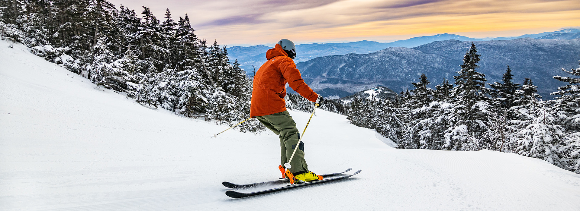 A skier in an orange coat and grey helmet with a view of surrounding mountains.