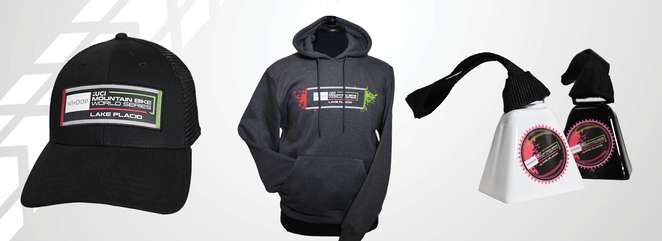 Grey background with white bike tire tracks on the left side and examples of UCI event branded merchandise from left to right including a black baseball style hat, a grey hoodie, and two cowbells in both white and black,
