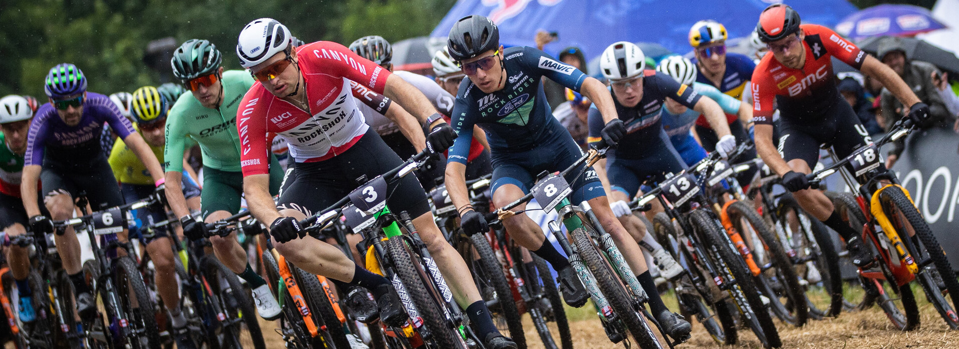 A large group of professional men mountain bikers in a race rounding a corner with focused looks on their faces.