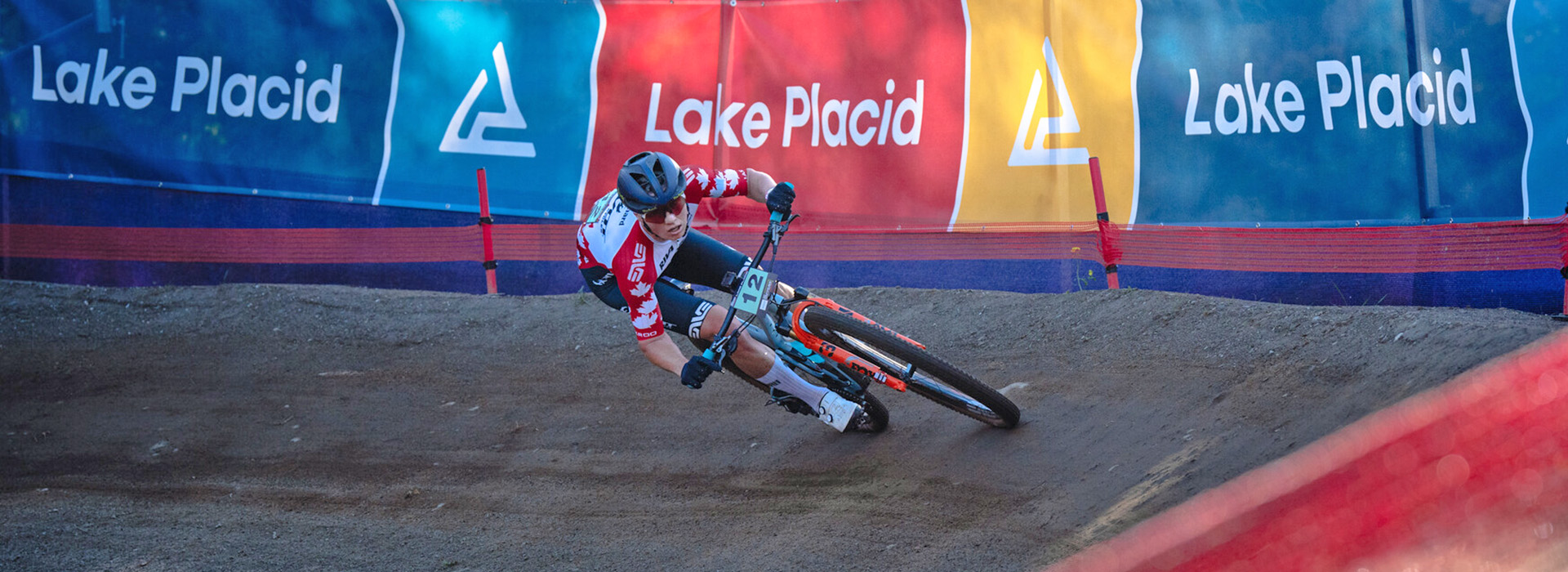 A female pro rider on the WHOOP UCI Mountain Bike World Series course at Mt Van Hoevenberg navigating a turn with Lake Placid banners in the background.