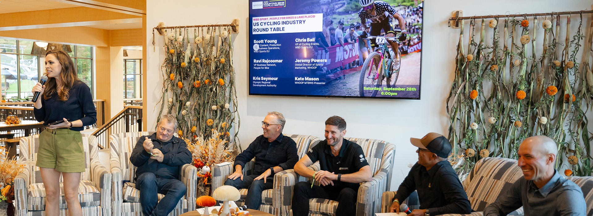 The roundtable panel in lounge chairs at Mt Van Hoevenberg with the moderator standing talking into a microphone and panelists seating and smiling.