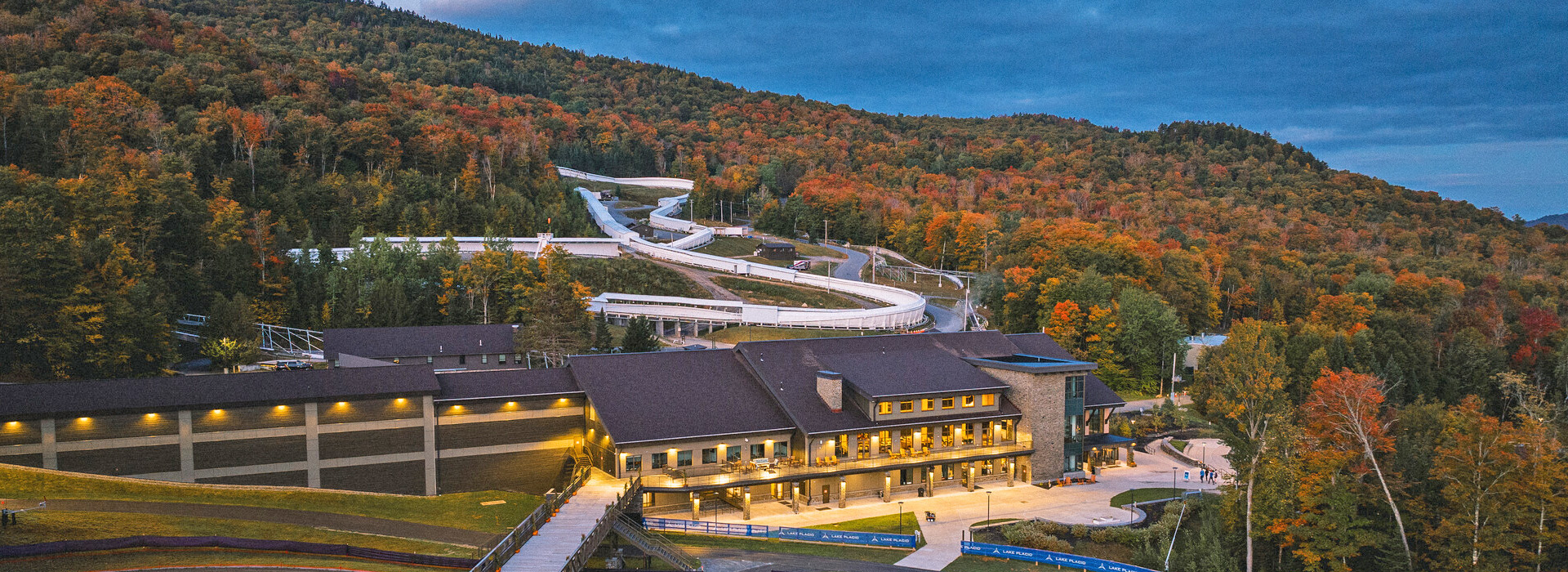 Drone shot of Mt Van Hoevenberg showing the Mountain Pass Lodge lit up near dusk with the fall forest and the combined sliding track on the mountain behind.