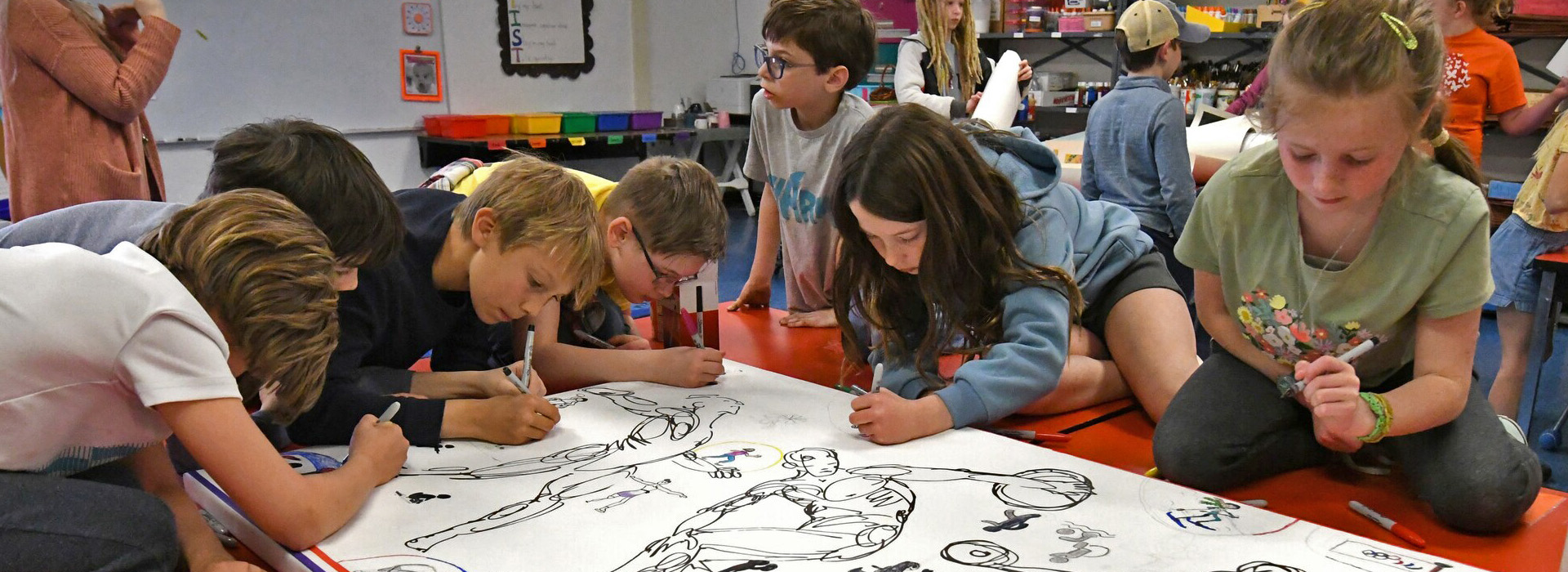 Seven students in a classroom leaning over and working on a drawing of athletes.