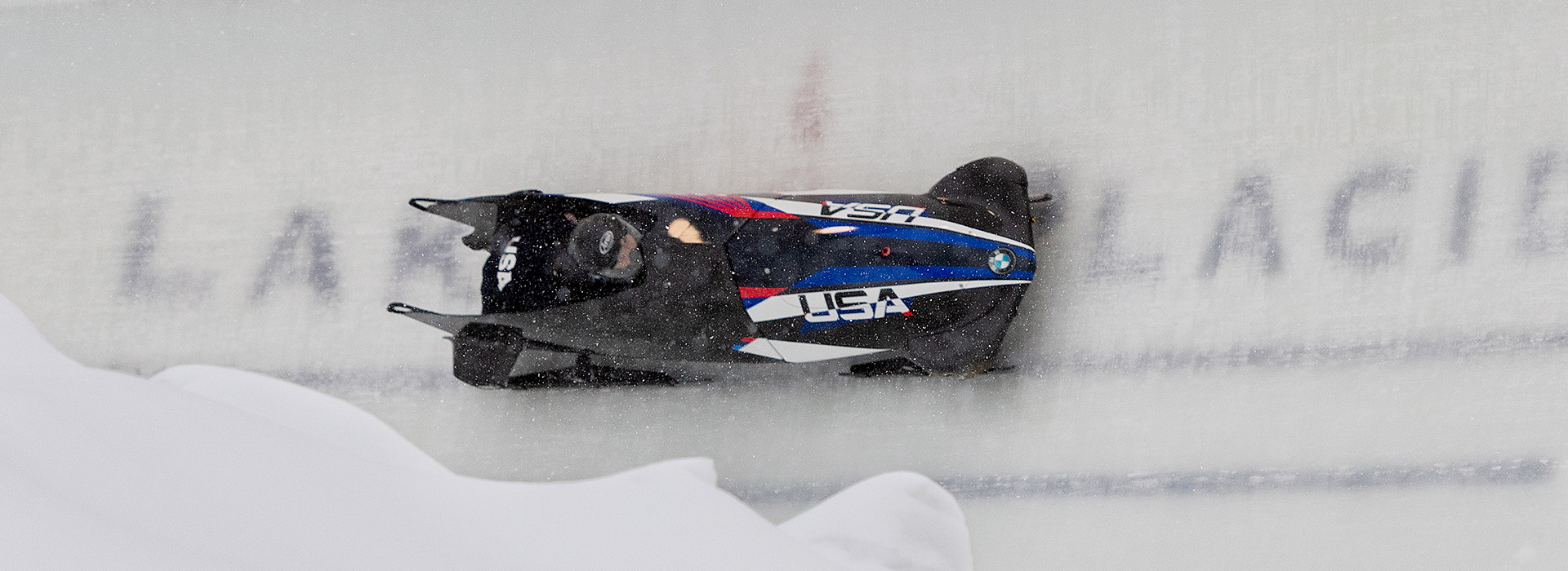 A two person Team USA bobsled races around a high banked turn on the Mt Van Hoevenberg combined track with the words "Lake Placid" imprinted in the ice under the sled.